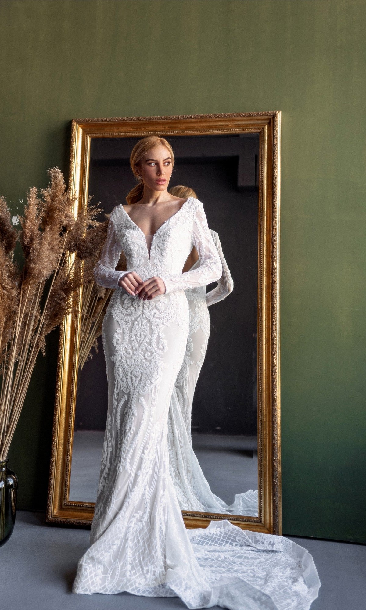 A woman in the Long Sleeve White Bridal Gown: Tina Holly BB021 stands before a large gold-framed mirror set against a green wall, with dried pampas grass arranged in a vase nearby.